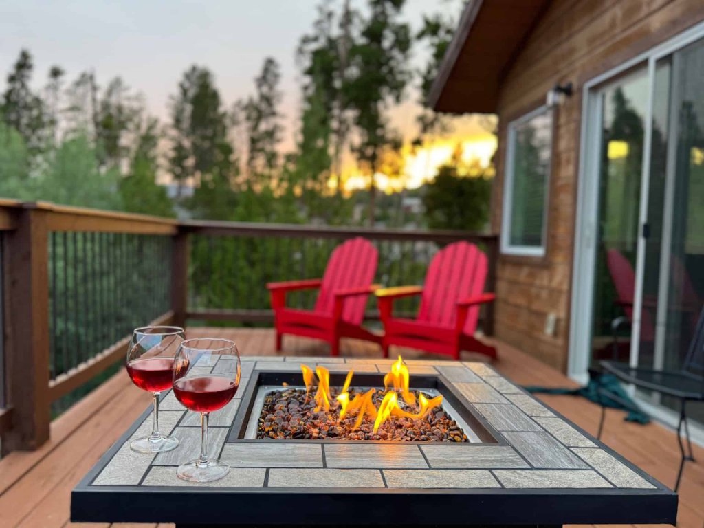 A cozy deck with a fire pit table, red Adirondack chairs, and wine glasses overlooking a forest at sunset in Grand County Colorado.