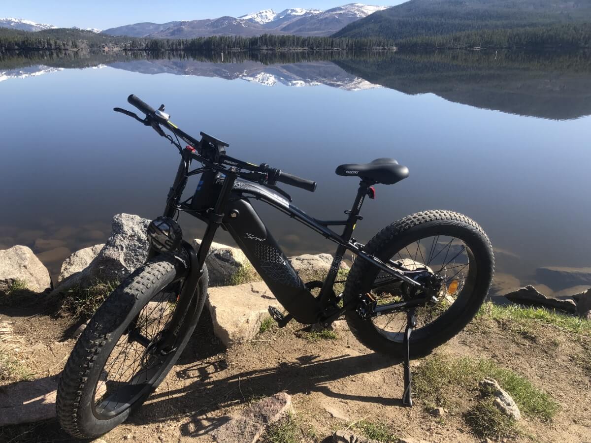 A black fat-tire mountain bike stands on a rocky shore beside a serene lake reflecting snow-capped mountains in Grand County Colorado.