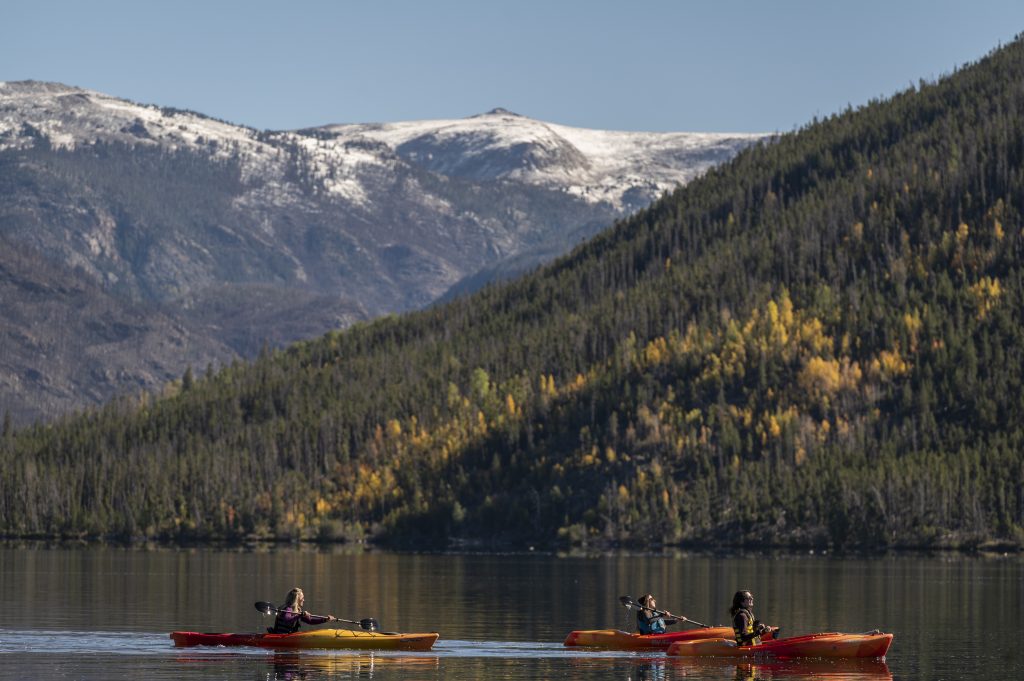 Three people kayaking on the water at Grand Lake Marina with forested mountains and snow-capped peaks in the background.