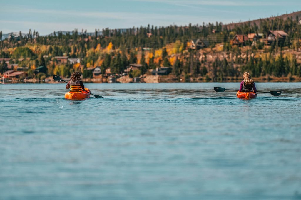 Two kayakers paddle on a calm lake surrounded by autumn trees and distant houses in Grand County Colorado.