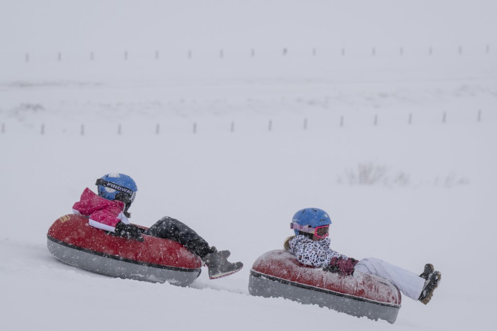 Two kids tubing side-by-side down a snowy hill in winter gear.