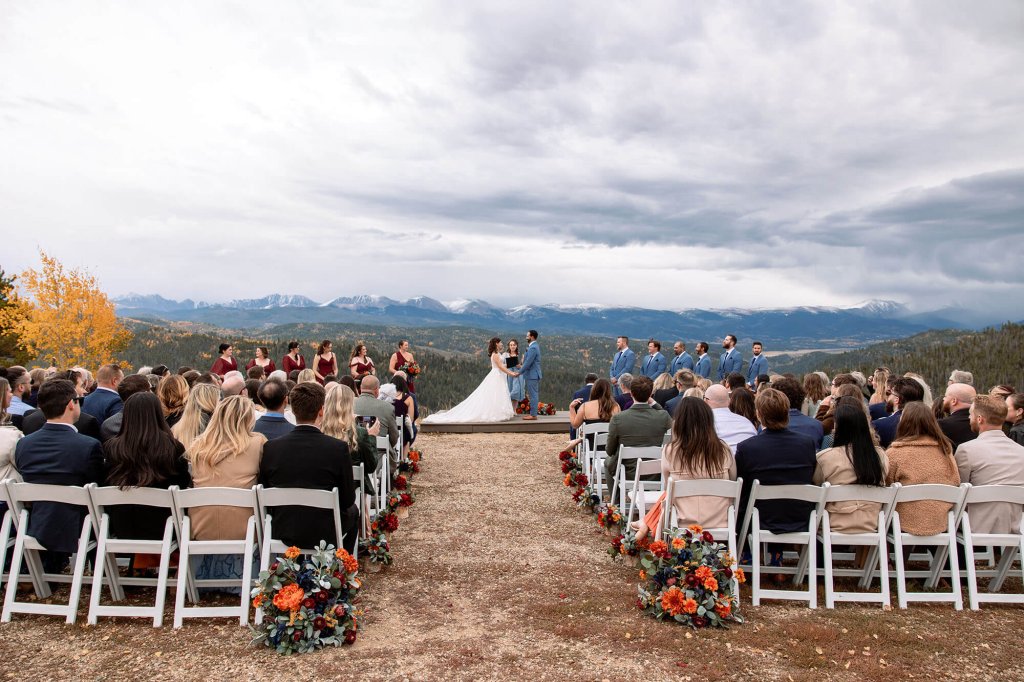 A couple exchanges vows on a mountain top with a breathtaking view of the Rocky Mountains in the background, surrounded by their wedding party and guests seated on white chairs at Granby Ranch in Grand County Colorado.