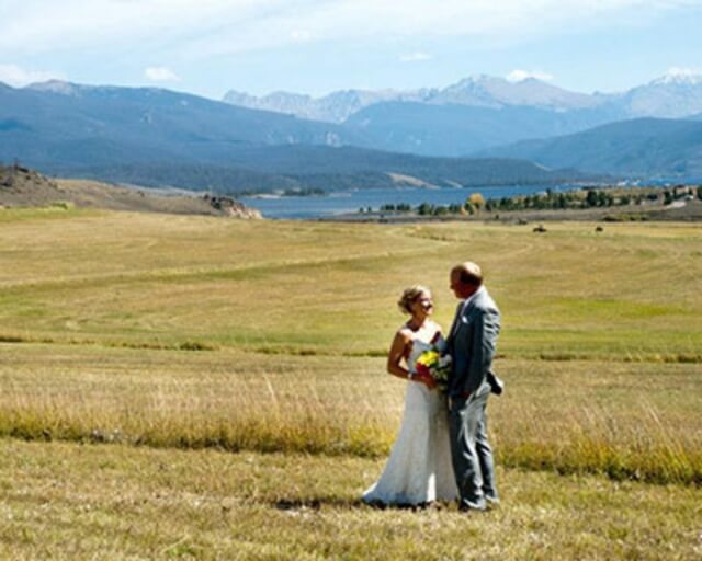 A bride and groom stand in a grassy field, with mountains and a lake in the distance in Grand County Colorado.