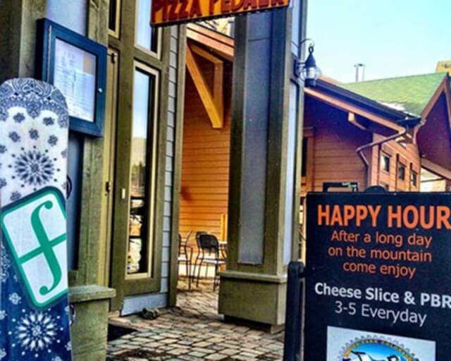 A snowboard leans against a building with a sign advertising a happy hour with cheese slices and PBR in Grand County Colorado.