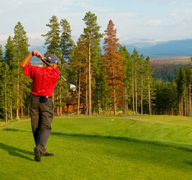 A golfer in a red shirt swings a club on a lush green course surrounded by tall trees in Grand County Colorado.