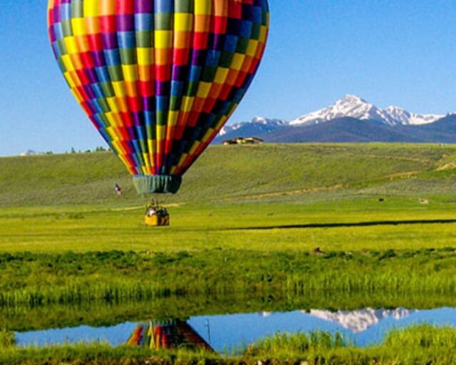 A colorful hot air balloon hovers above a serene pond with mountain peaks in the distance in Grand County Colorado.