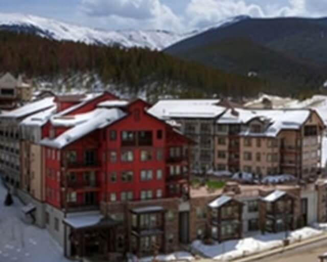 A cluster of red and beige buildings with snow-covered roofs nestled among evergreen trees at the base of snow-capped mountains in Grand County Colorado.