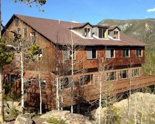 A rustic wooden cabin with multiple stories and a deck, nestled among trees on a rocky hillside in Grand County Colorado.