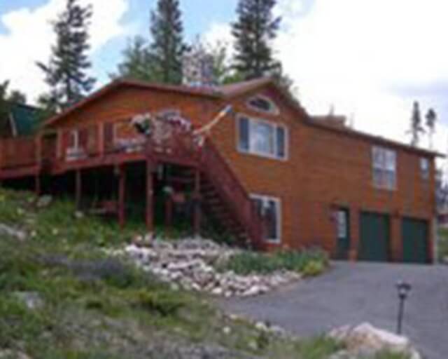 A rustic, two-story wooden house with a wrap-around porch and a staircase leading to the entrance in Grand County Colorado.