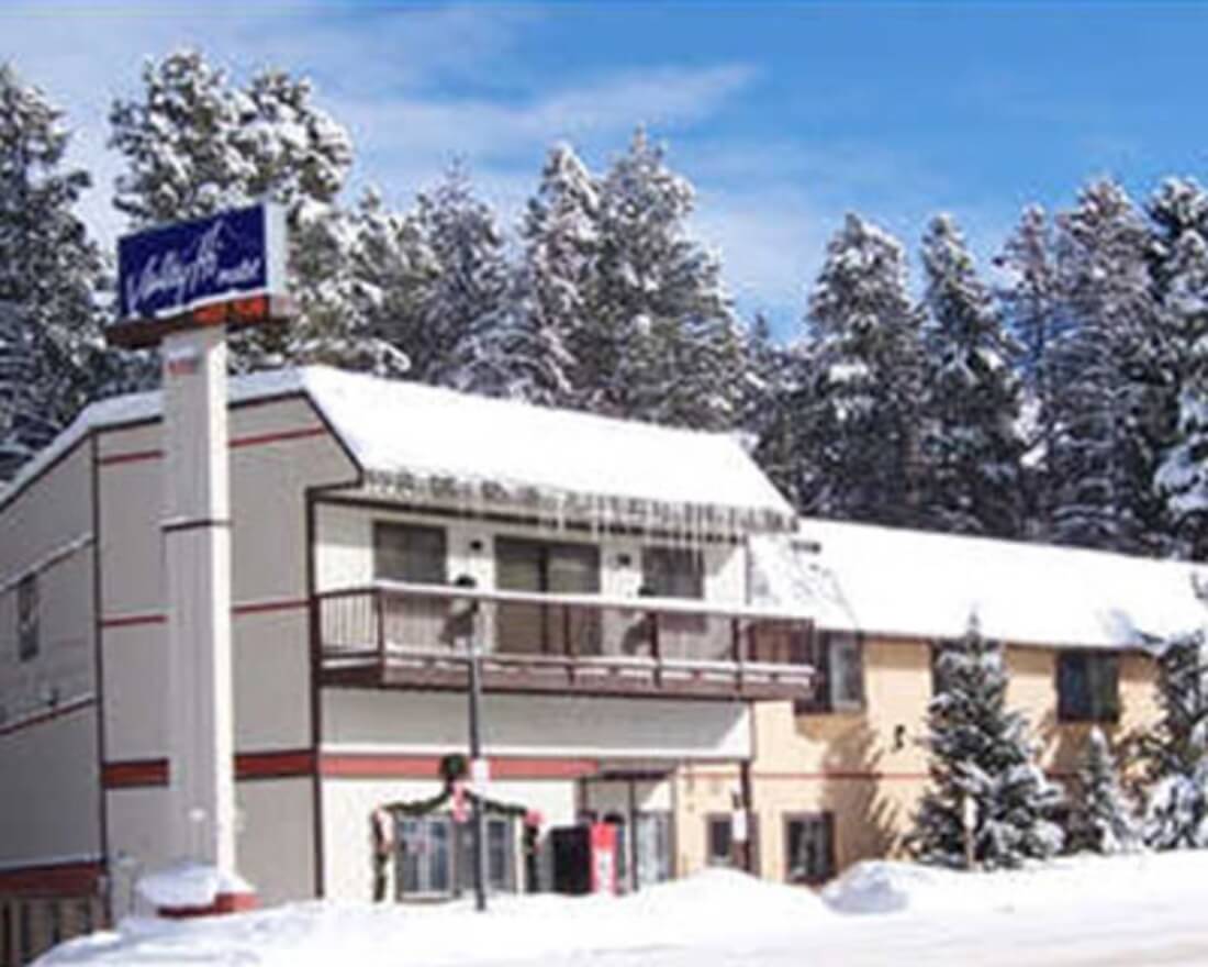 A two-story motel building with a snow-covered roof and balcony, surrounded by snow-laden evergreen trees in Grand County Colorado.
