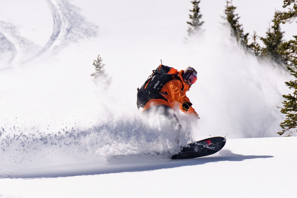 A snowboarder in an orange jacket and black backpack carves through deep snow, kicking up a cloud of powder in Grand County Colorado.
