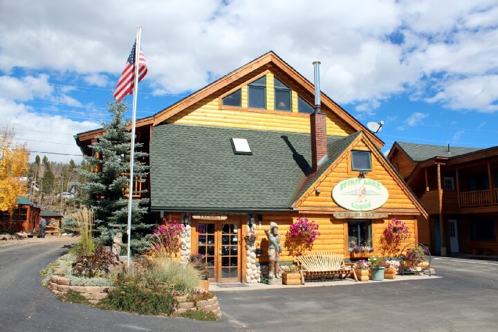 A charming log cabin with a steep roof, surrounded by flowers and an American flag in Grand County Colorado.
