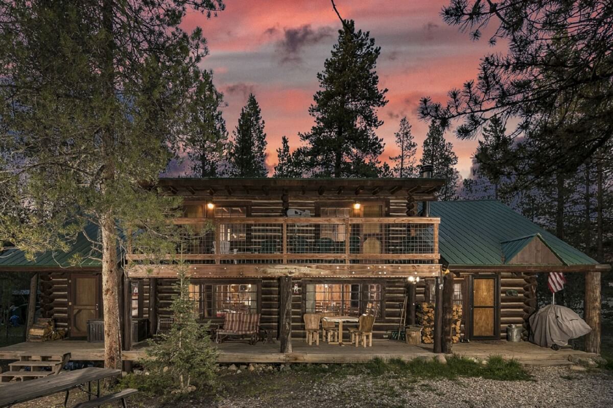 Rustic log cabin with wrap-around porch and stone fireplace, nestled among tall pine trees in Grand County Colorado.