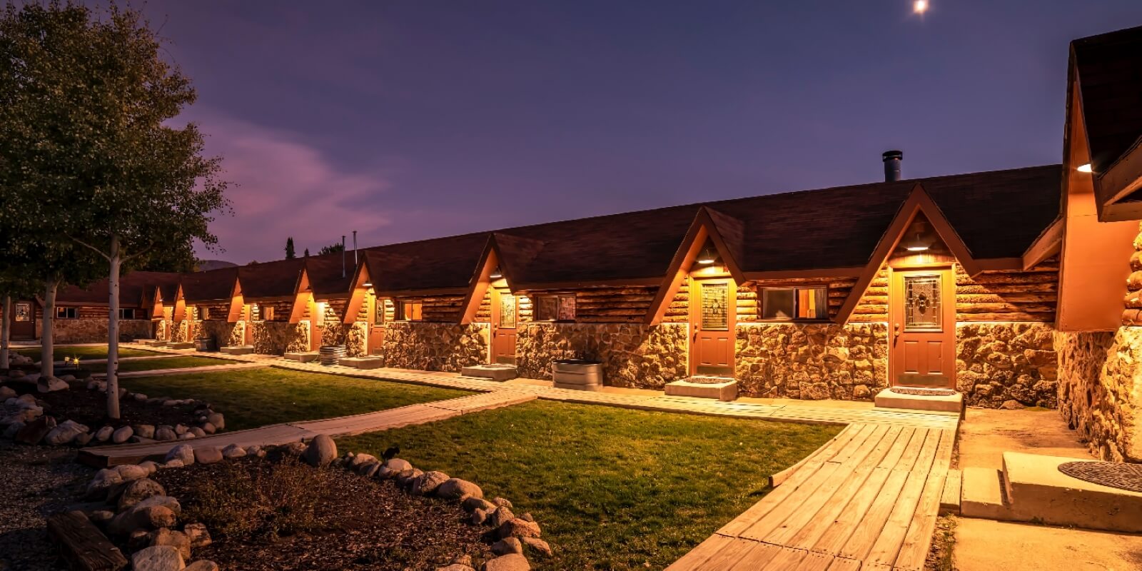 A row of rustic log cabins with stone foundations, illuminated by warm lights at dusk in Grand County Colorado.