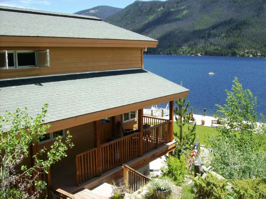 A rustic wooden cabin with a porch overlooks a serene lake surrounded by mountains and trees in Grand County Colorado.