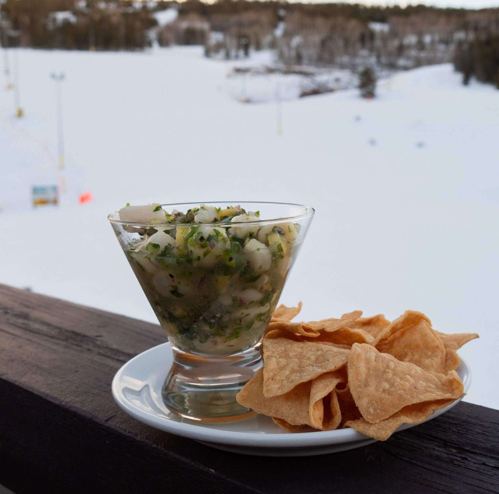 A glass filled with a vibrant green salsa or ceviche sits on a wooden railing, accompanied by a plate of crispy tortilla chips, with a snowy mountain landscape in the background in Grand County Colorado.