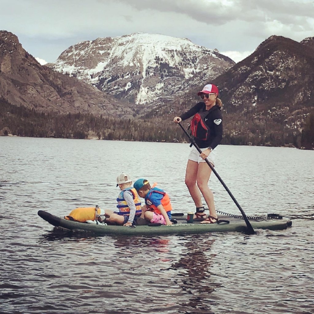 A woman paddles a stand-up board with three children on a lake surrounded by mountains in Grand County Colorado.