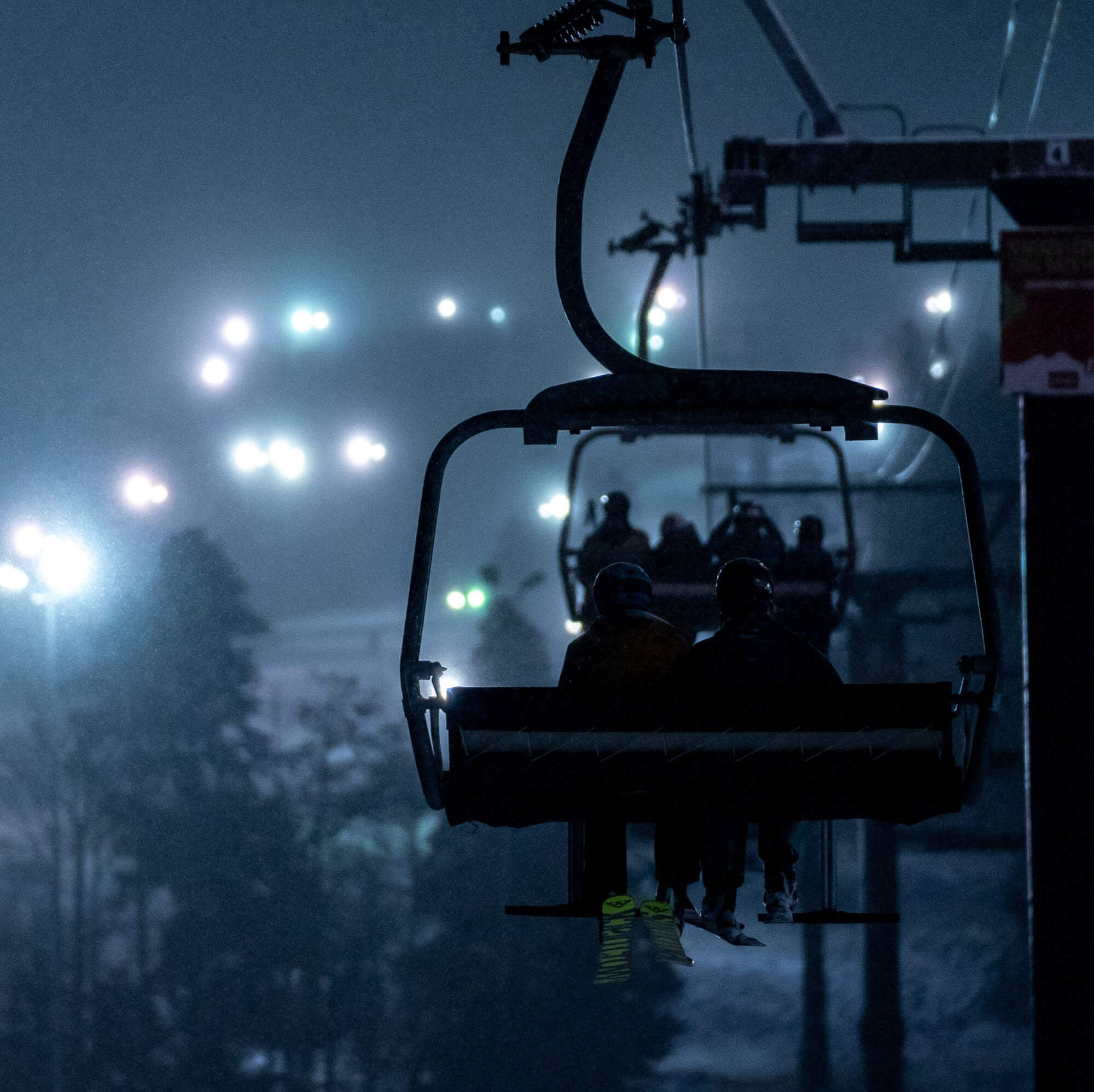 Skiers ride a chairlift at night, silhouetted against a backdrop of twinkling lights and snow-covered trees in Grand County Colorado.