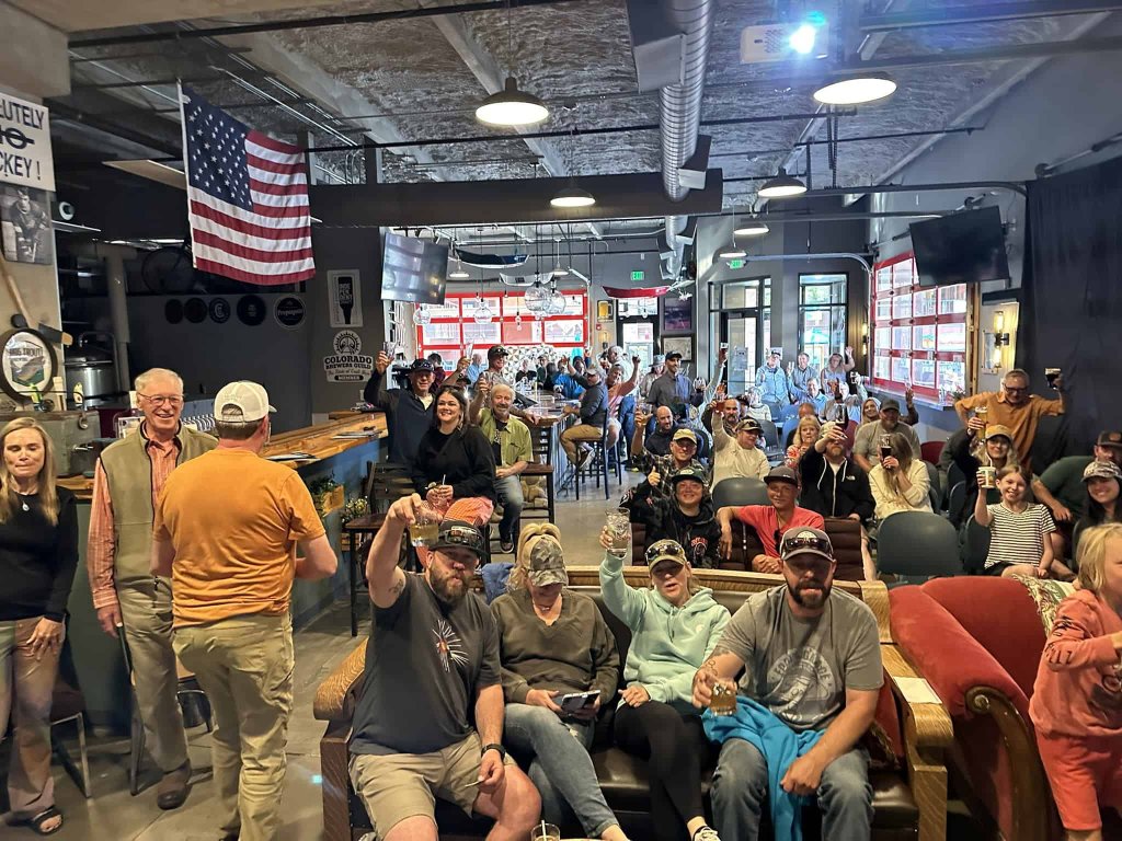 Crowded bar with people raising glasses, American flag, and exposed ceiling beams in Grand County Colorado.