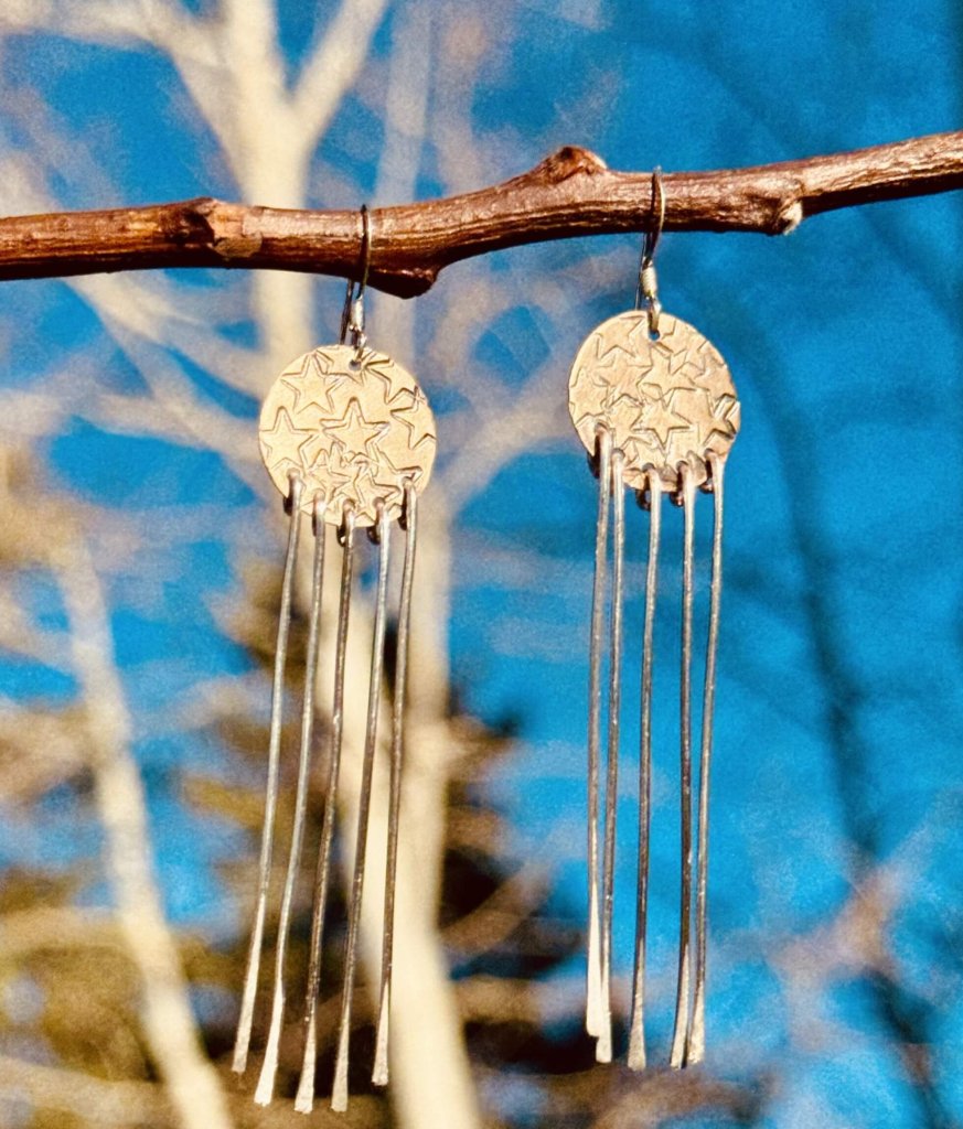 A pair of star-shaped earrings with long, thin metal strands hanging from them, suspended from a tree branch against a clear blue sky in Grand County Colorado.