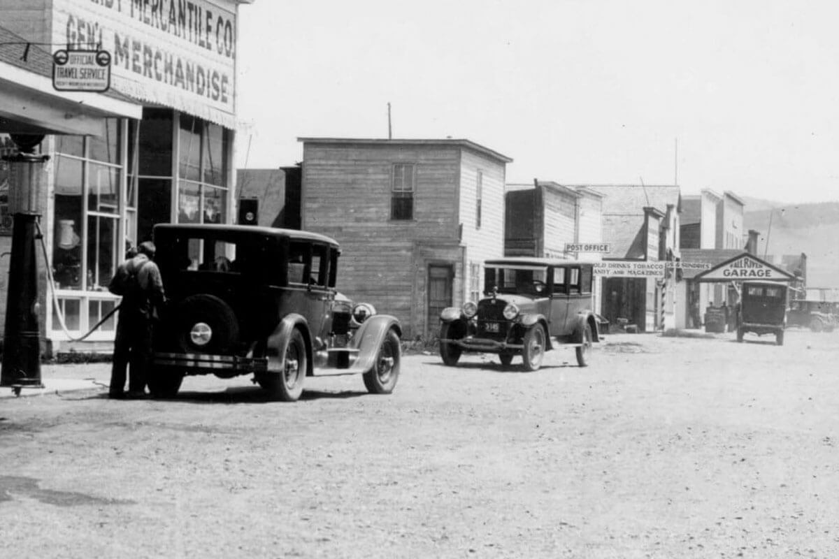 A vintage black-and-white photograph of a small town street scene with early 20th-century automobiles and storefronts in Grand County Colorado.