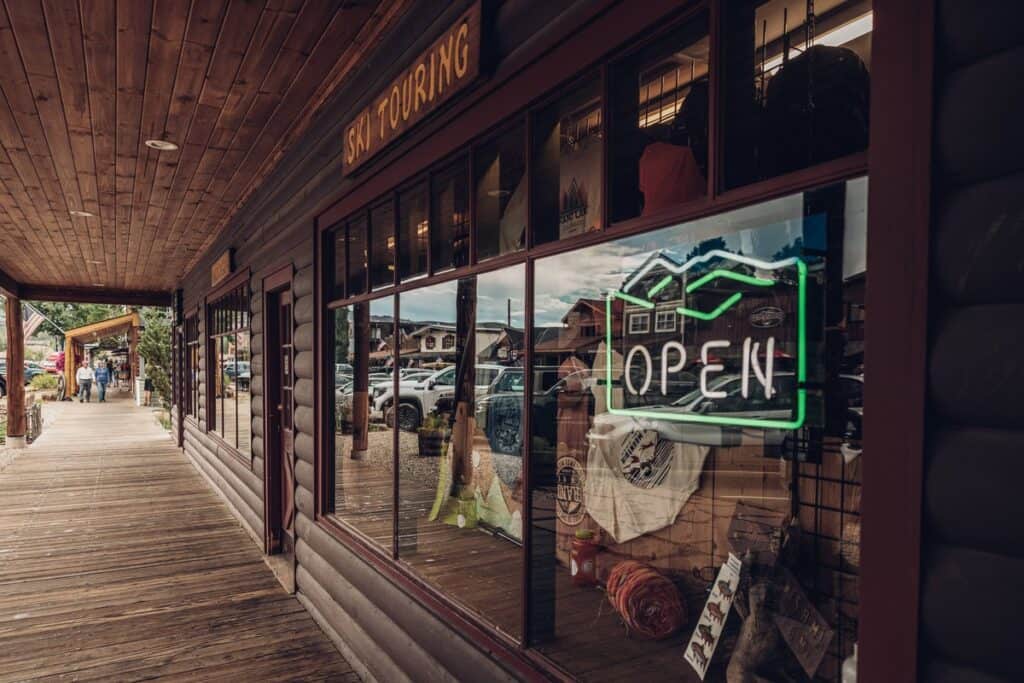 A wooden storefront with a row of windows has a lit "Open" sign. The reflection shows parked cars and buildings across the street. A glimpse of the store’s interior is visible through the windows.