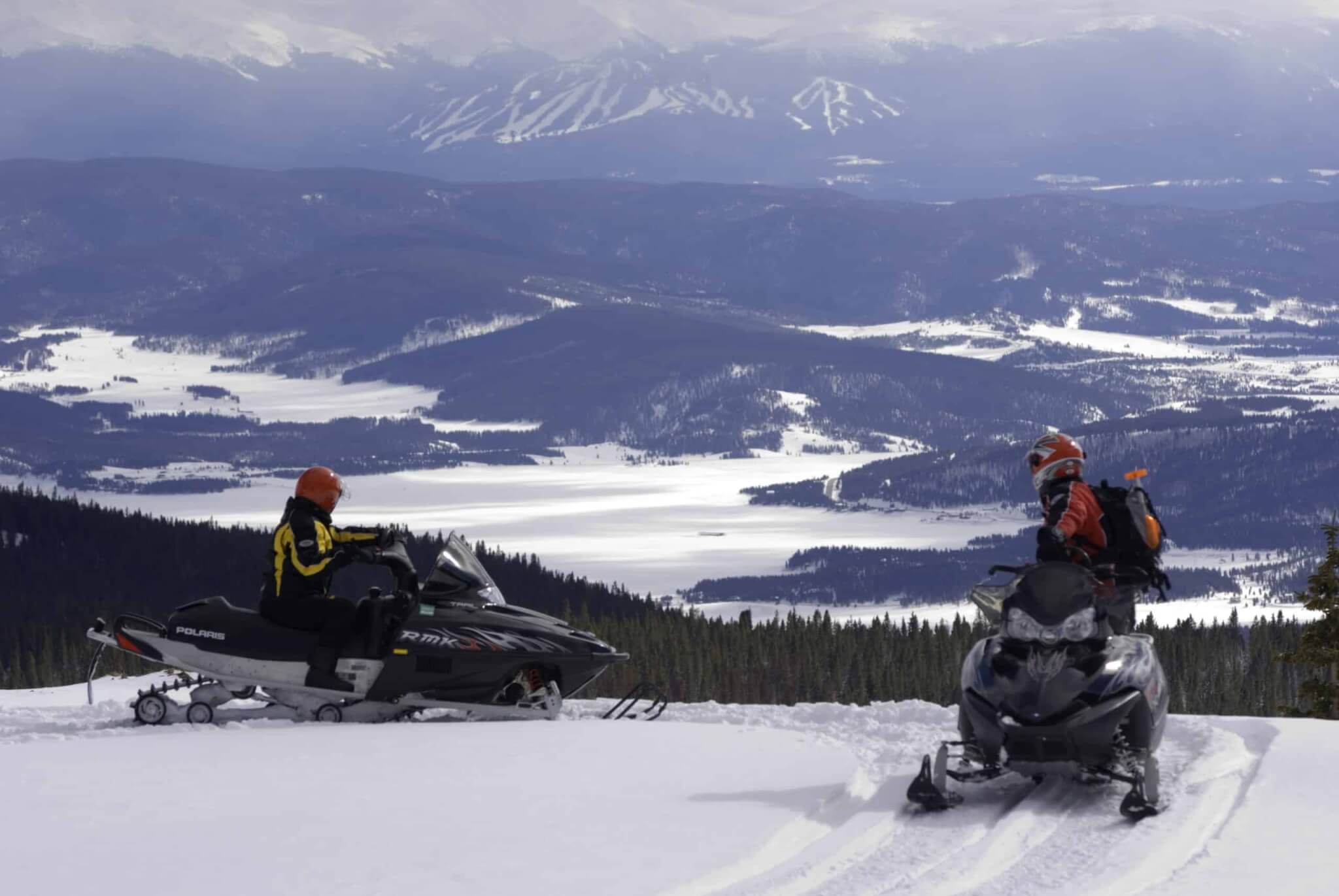 Two snowmobilers on a snowy mountain ridge overlooking a vast, snow-covered valley and distant mountains in Grand County Colorado.