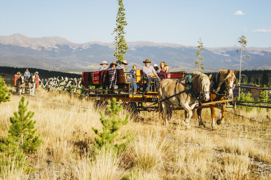 A horse-drawn wagon filled with people travels through a grassy field with mountains in the background in Grand County Colorado.