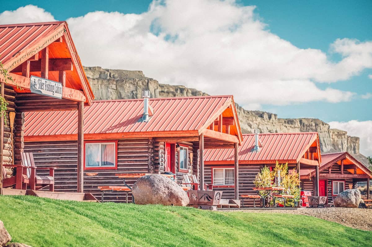 Row of charming wooden log cabins with red roofs, under a clear blue sky, set in a lush green landscape in Kremmling, Colorado.