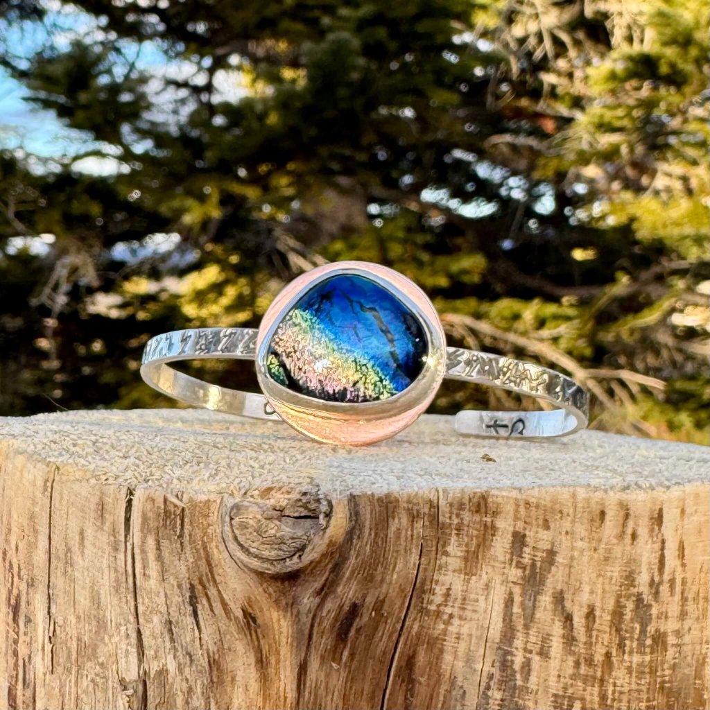A handcrafted silver cuff bracelet with a large, iridescent blue and green stone set in the center, resting on a rustic wooden stump against a backdrop of evergreen trees in Grand County Colorado.