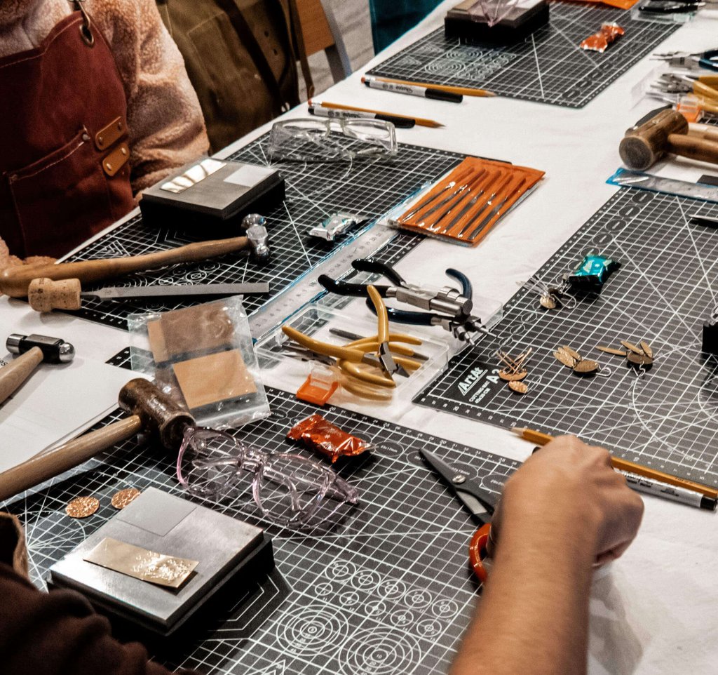 A detailed view of a crafting table filled with various tools and materials, including hammers, pliers, scissors, and metalworking supplies, with hands actively engaged in creating jewelry in Grand County Colorado.