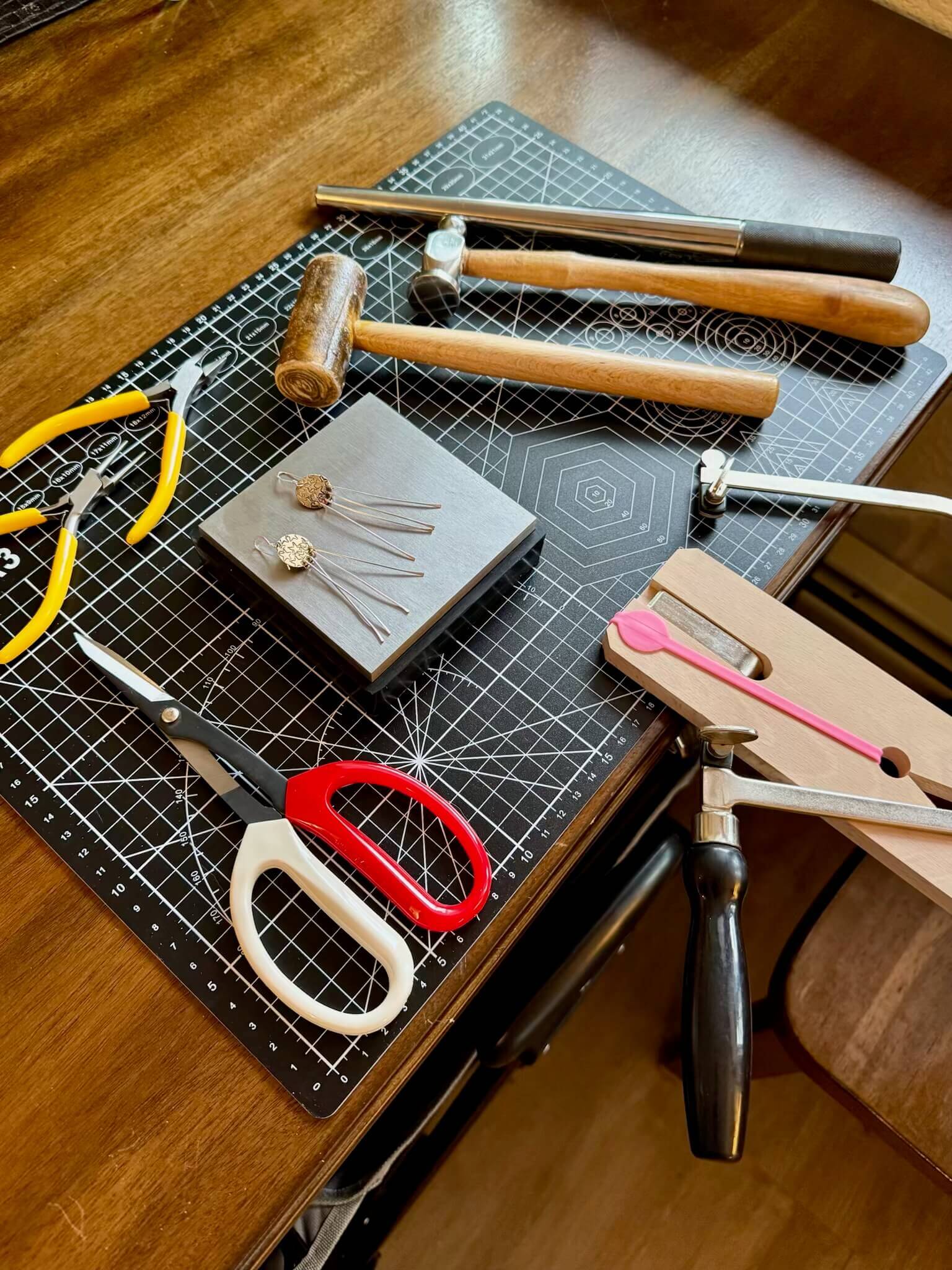A variety of jewelry-making tools and materials are neatly arranged on a black and white grid cutting mat, including pliers, hammers, a mallet, scissors, and metal stamping tools in Grand County Colorado.