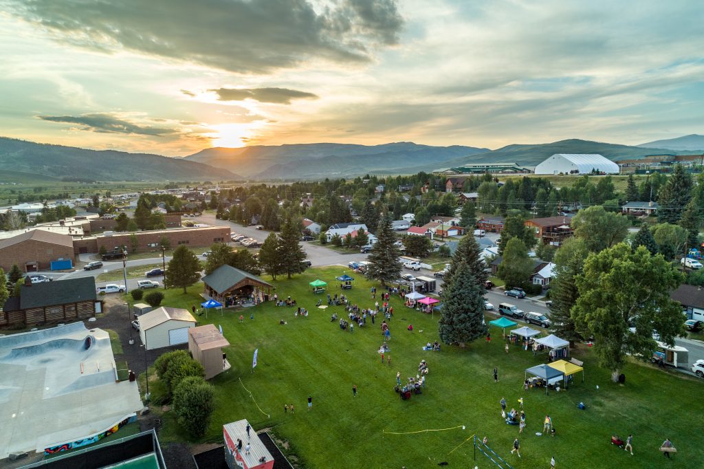 Aerial view of a community event in a park at sunset with tents, people, and mountains in the background.