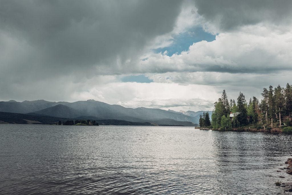 Granby Lake with forested shoreline, distant mountains, and storm clouds overhead.