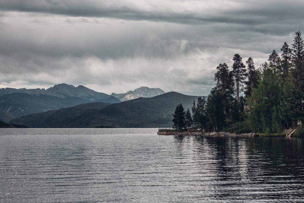 Lake with forested shoreline and mountains in the distance under cloudy skies.