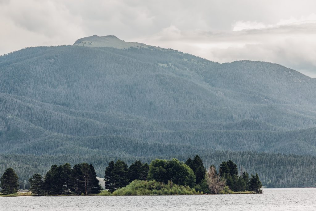 A serene lake with lush green trees on an island, surrounded by rolling mountains and a cloudy sky in Grand County Colorado.