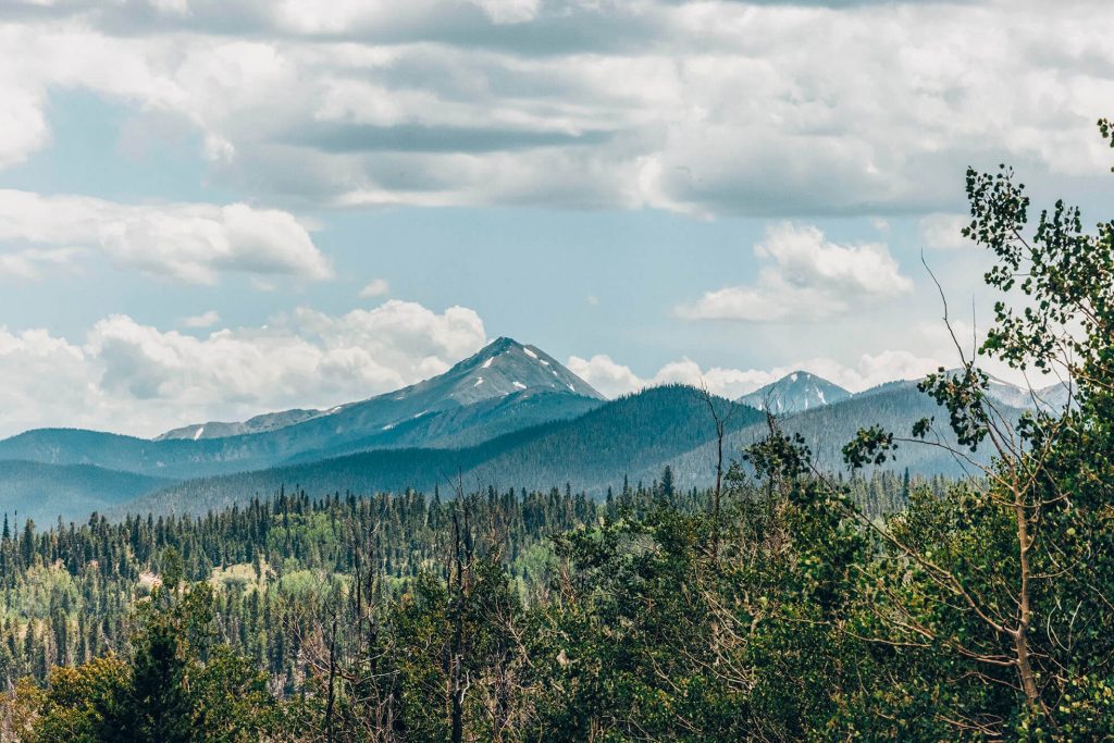 A vast mountain range with snow-capped peaks rises above a lush green forest under a cloudy sky in Grand County Colorado.