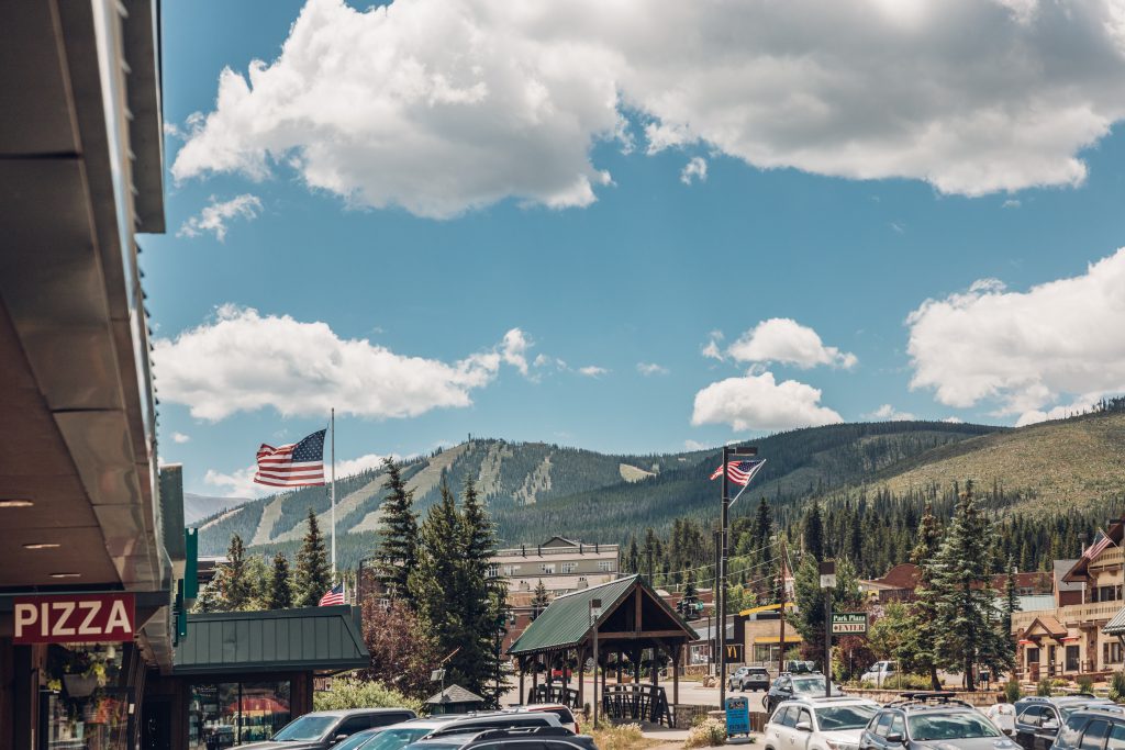 Downtown Winter Park with American flags, shops, and cars, set against a backdrop of forested mountains and a partly cloudy sky.