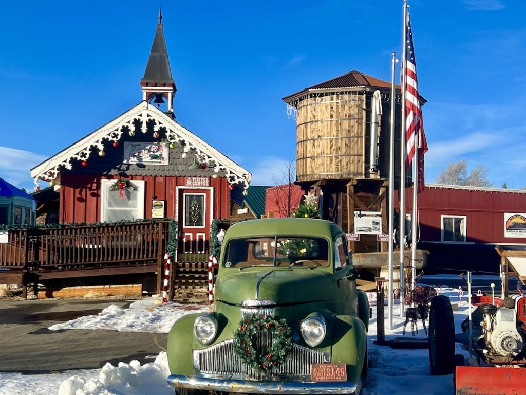 Vintage green truck with wreath parked in snow near red barn-like building with steeple in Grand County Colorado.