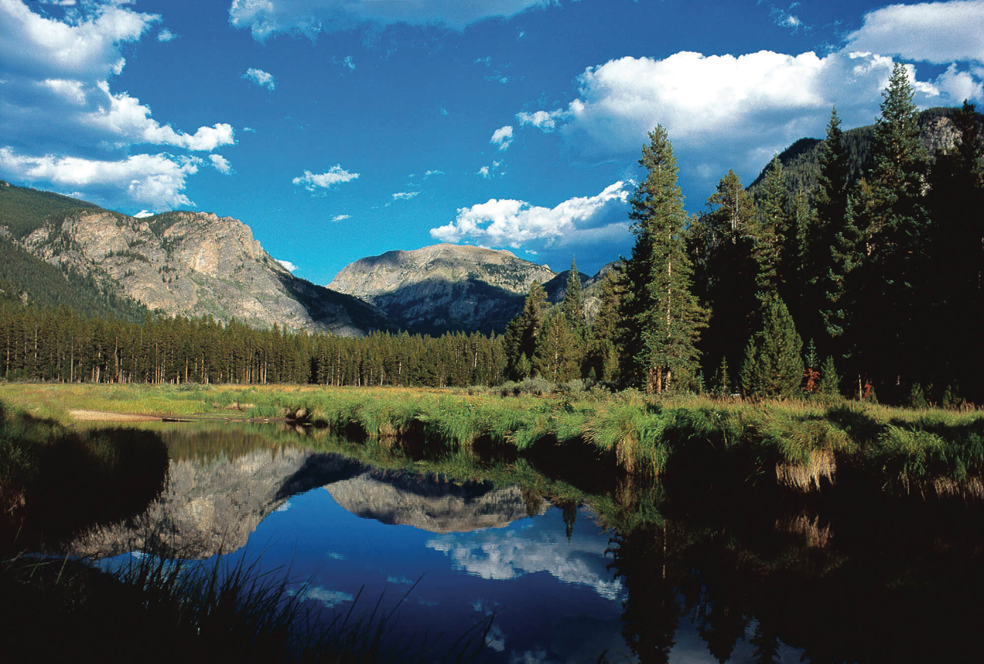 A serene mountain landscape with a calm lake reflecting the surrounding peaks and forests under a bright blue sky with fluffy white clouds in Grand County Colorado.