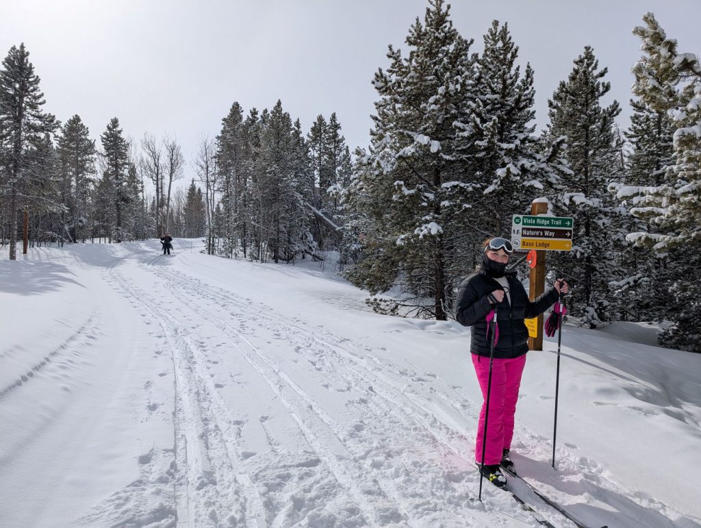 A skier stands on a snow-covered trail, surrounded by tall pine trees and a signpost in Grand County Colorado.