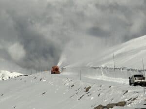 A snowplow clears a snow-covered mountain road with high snowbanks on either side. A pickup truck is parked to the right of the road, and clouds fill the sky in the background.