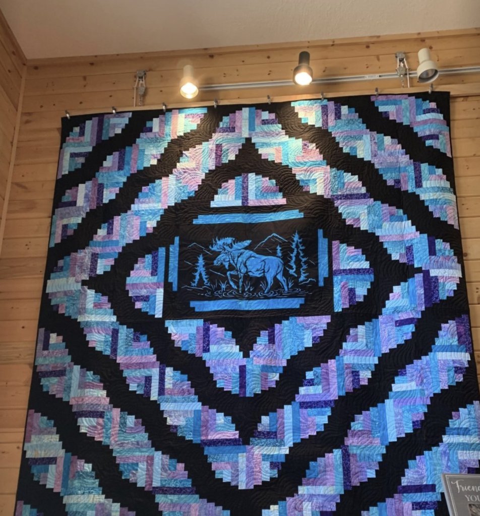 A large, intricately pieced quilt with a moose and trees in the center, hung on a wooden wall in Grand County Colorado.