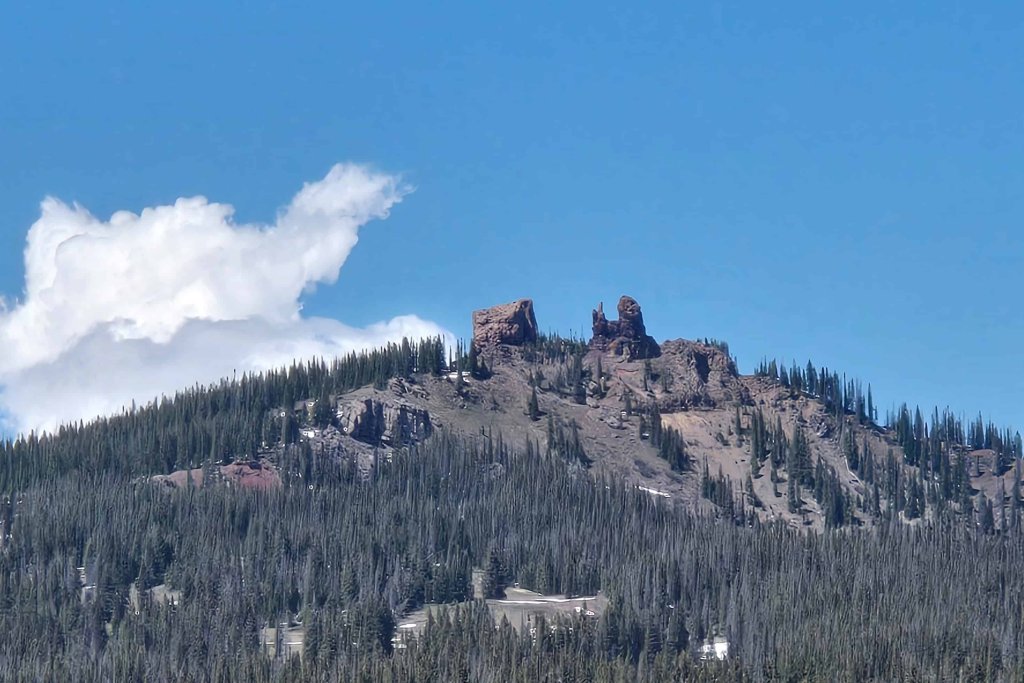 A rocky mountain peak rises above a dense evergreen forest under a blue sky with a fluffy white cloud in Grand County Colorado.