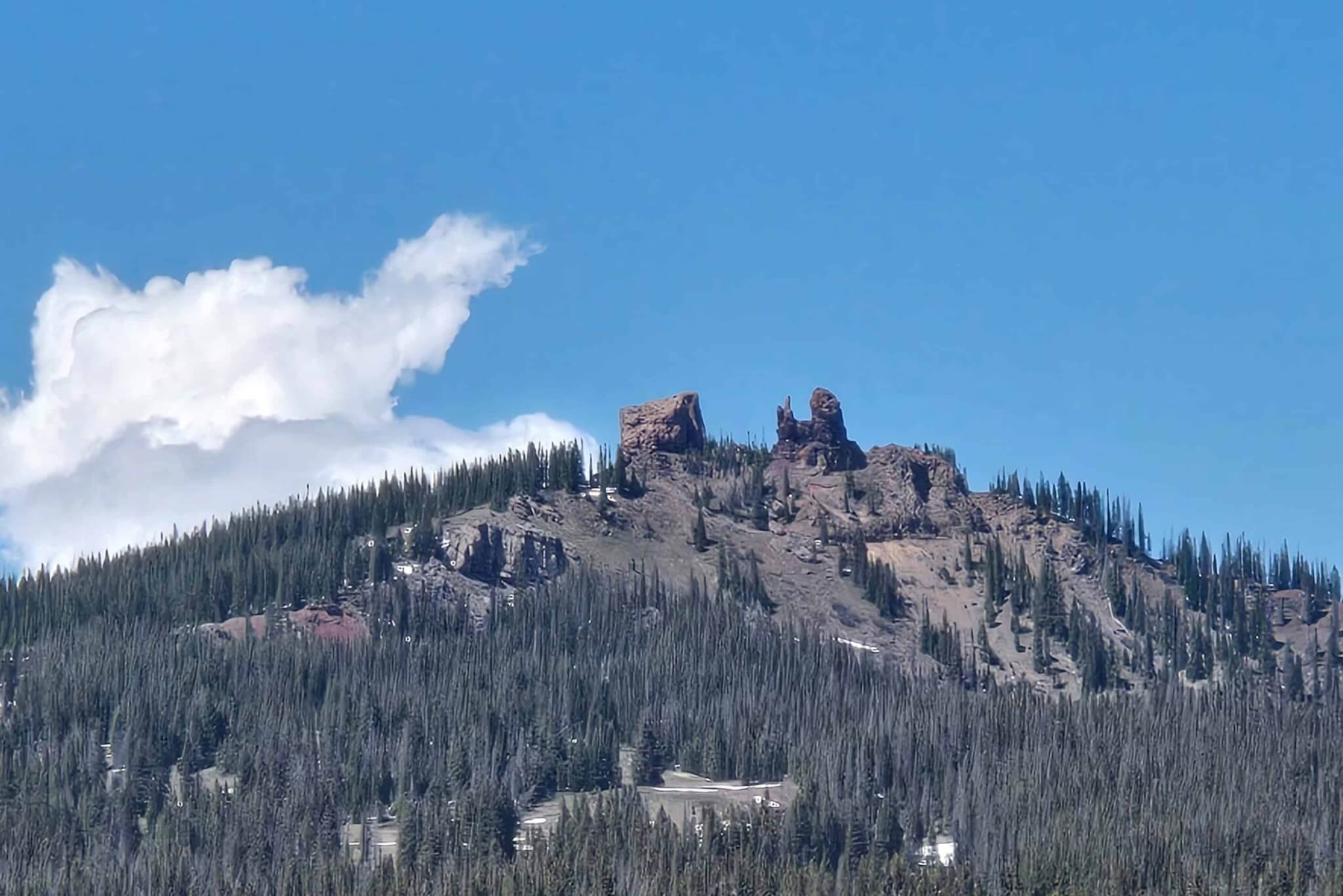 A rocky mountain peak rises above a dense evergreen forest under a blue sky with a fluffy white cloud in Grand County Colorado.