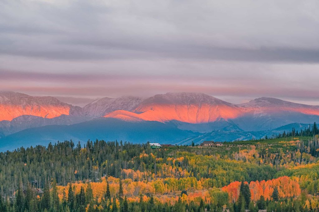 Sunrise paints the snow-capped mountains with warm hues, above a forest of autumn colors in Grand County Colorado.