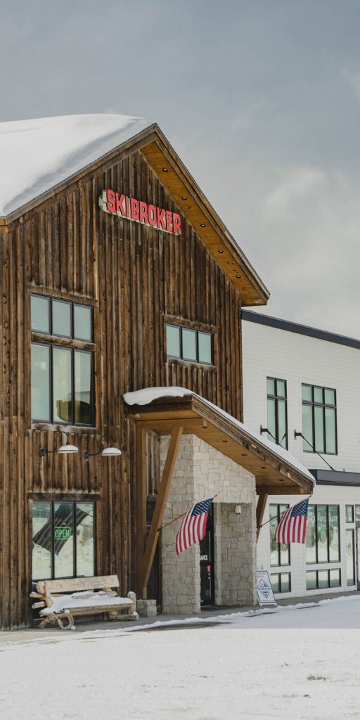 A rustic wooden building with a snow-covered roof, featuring a sign that reads 'SKIBROKER' and American flags hanging from the entrance in Grand County Colorado.