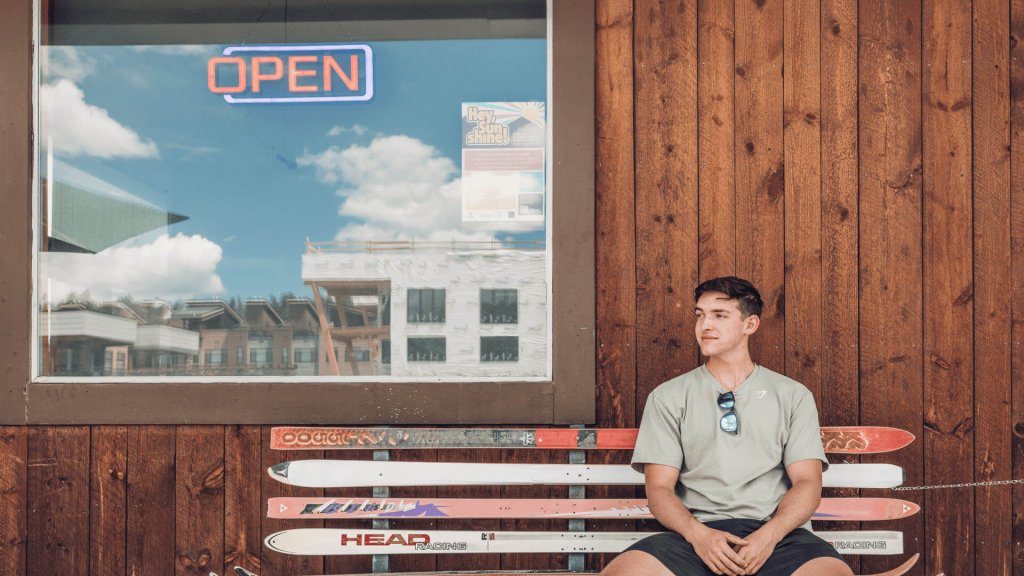 A young man sits on a bench made of colorful skis, wearing sunglasses and a casual t-shirt, with a wooden building and an 'OPEN' sign in the background in Grand County Colorado.