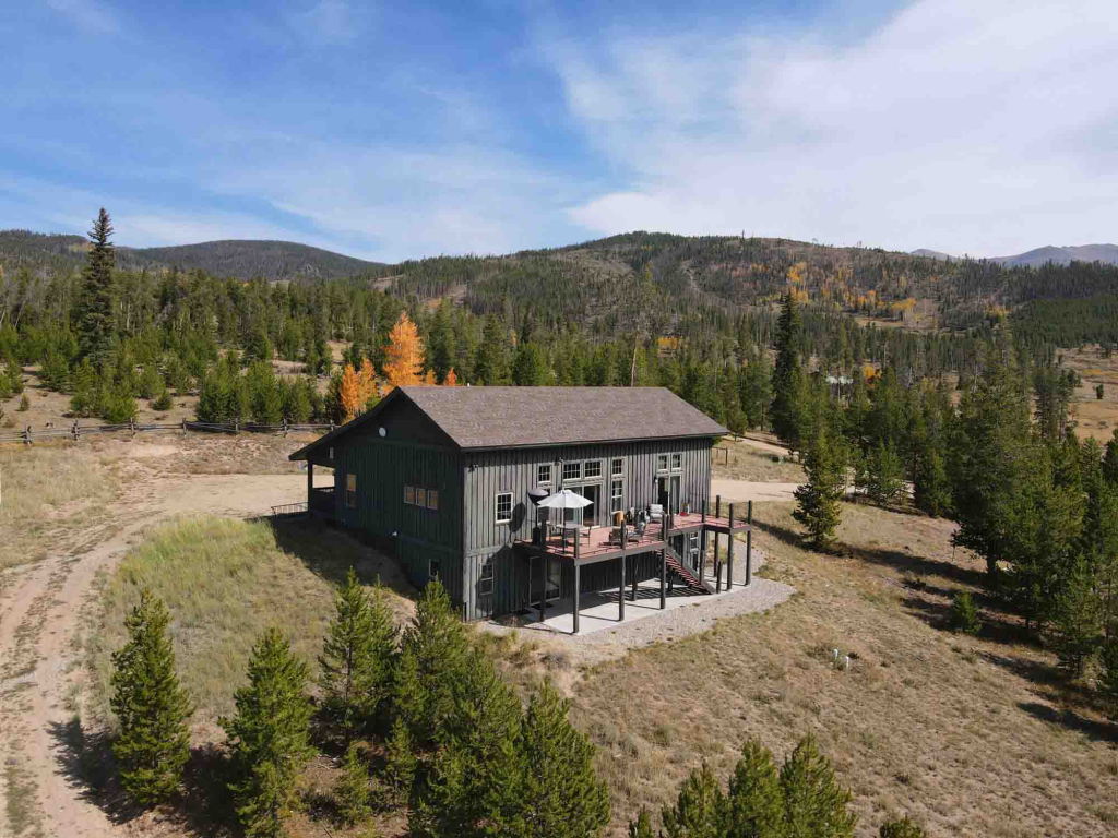 A rustic wooden cabin with a deck sits on a hill surrounded by trees and mountains in Grand County Colorado.