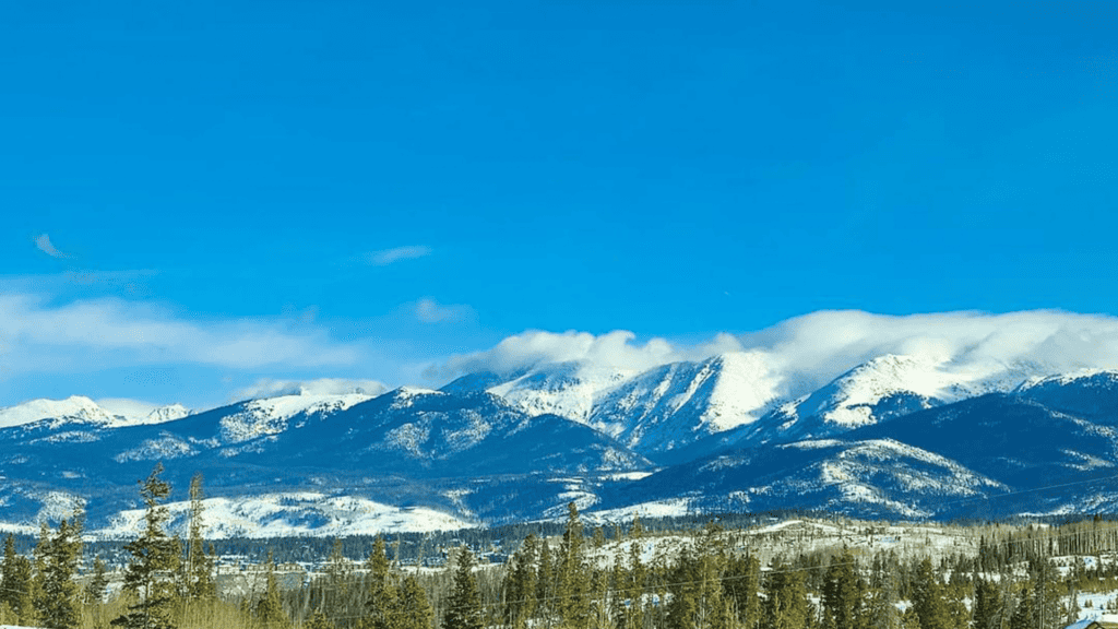 Snow-covered mountain peaks in Grand County, Colorado, with a clear blue sky and evergreen trees in the foreground, creating a stunning winter landscape.